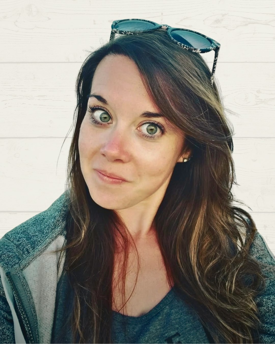 A woman with long brown hair and green eyes, wearing sunglasses on her head, a dark gray shirt, and a hoodie, standing in front of a white wooden background.