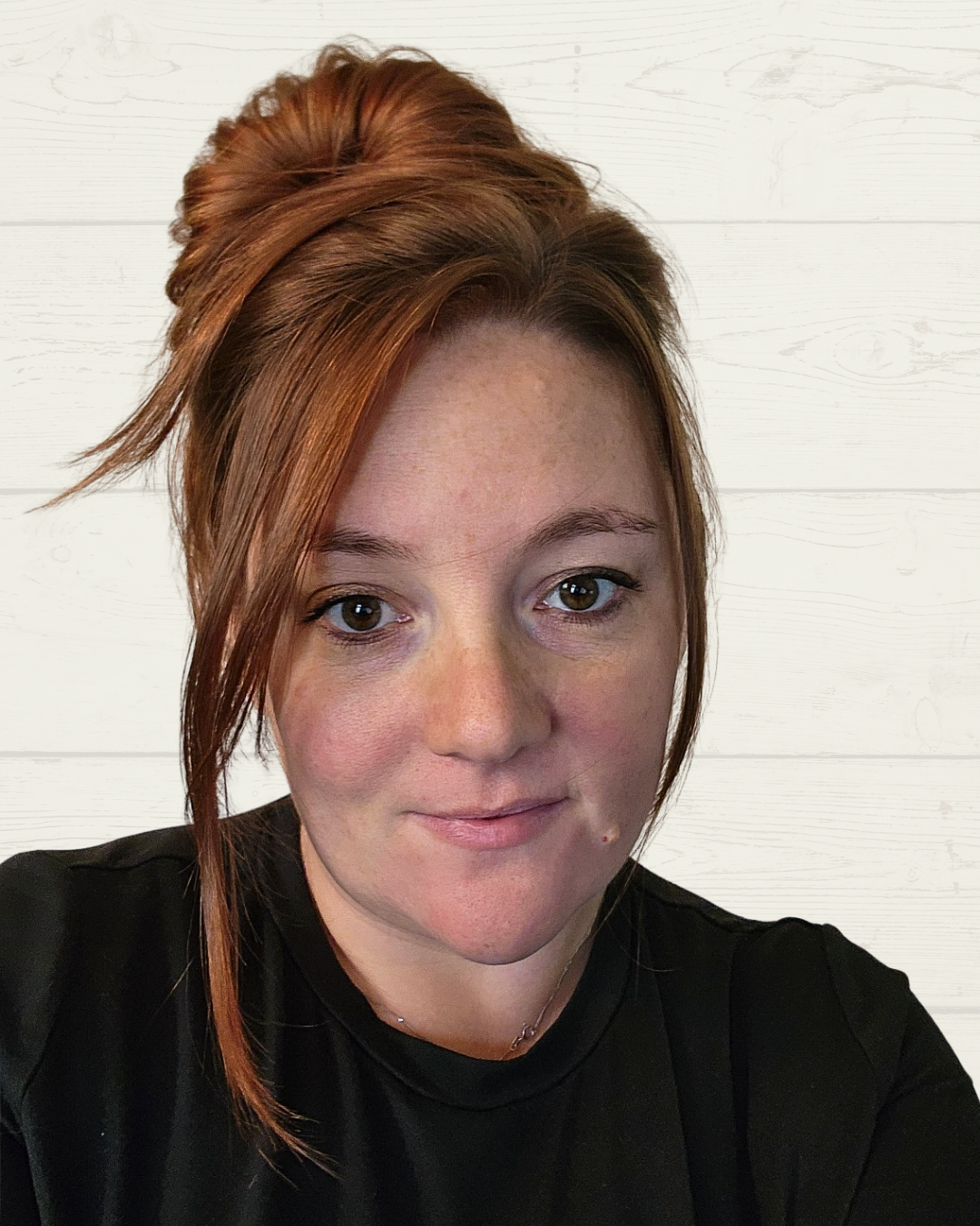 Close-up of a woman with red hair styled in an updo, wearing a black shirt, standing against a white wooden wall.