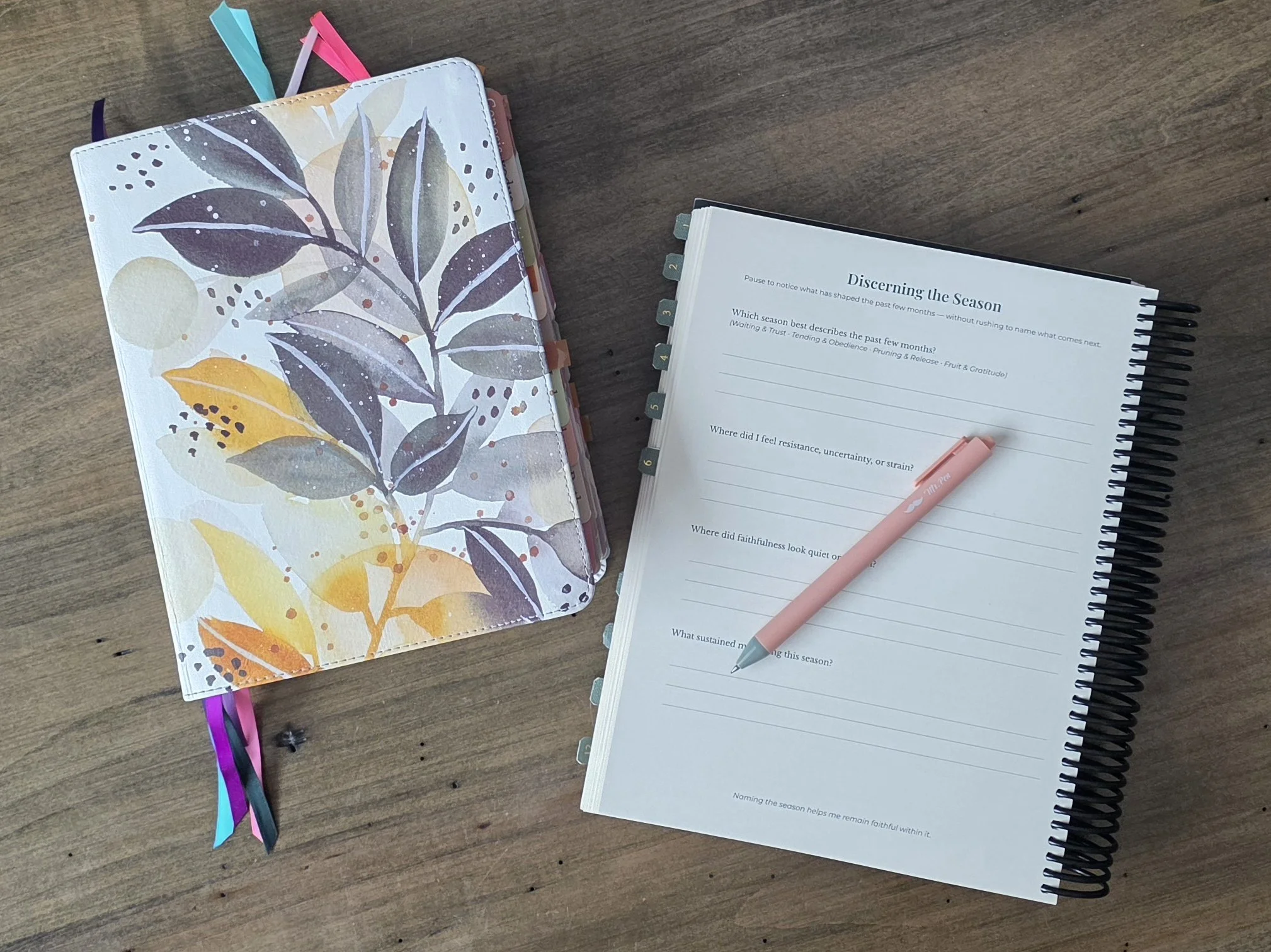 Open spiral Christian planner with pink pen next to bible with watercolor leaf design cover bible with colorful ribbon bookmarks on a wooden desk.