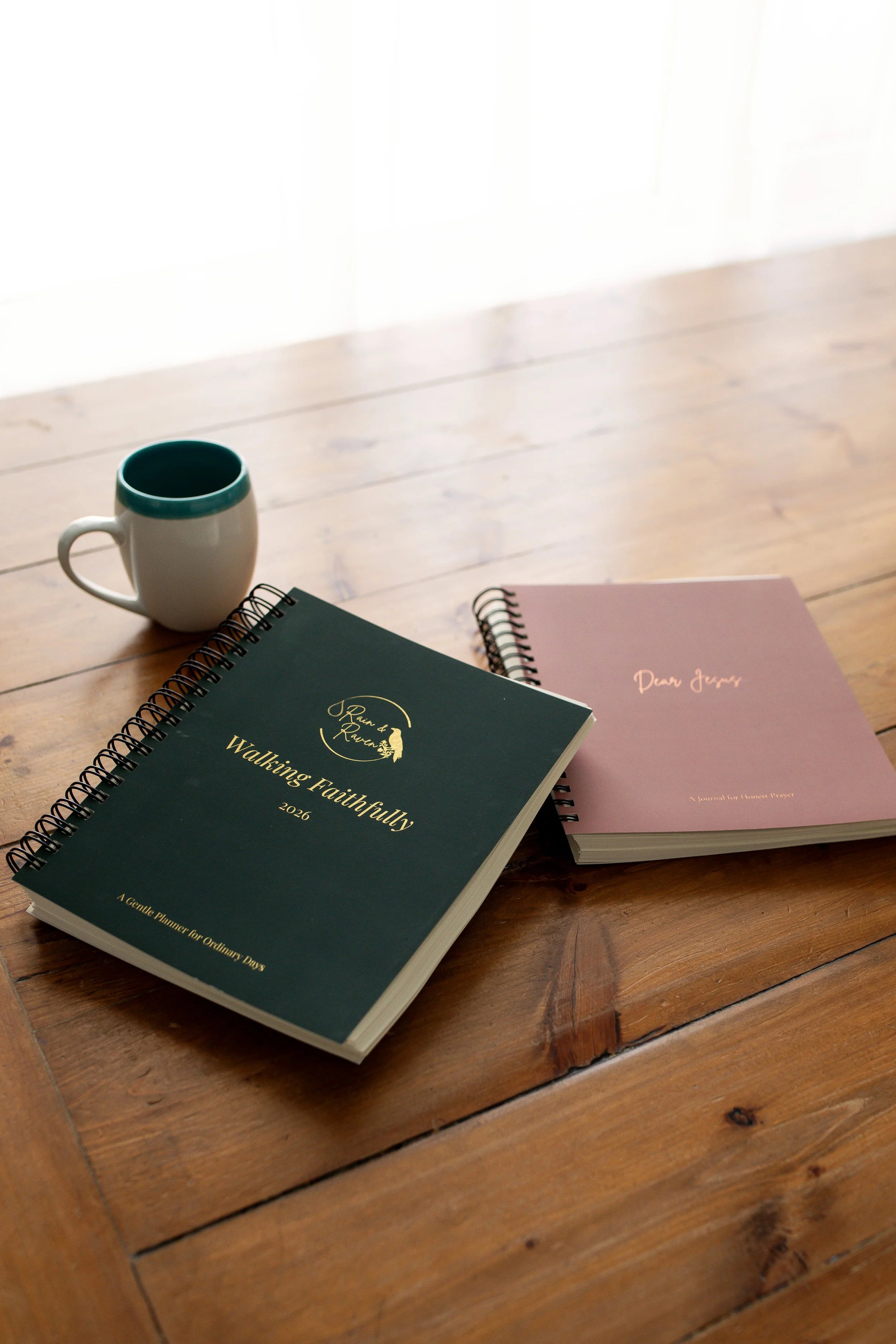 A wooden table with a white mug with a teal interior, a green  Christian planner labeled 'Walking Faithfully 2026,' and a pink journal labeled 'Dear Jesus'.