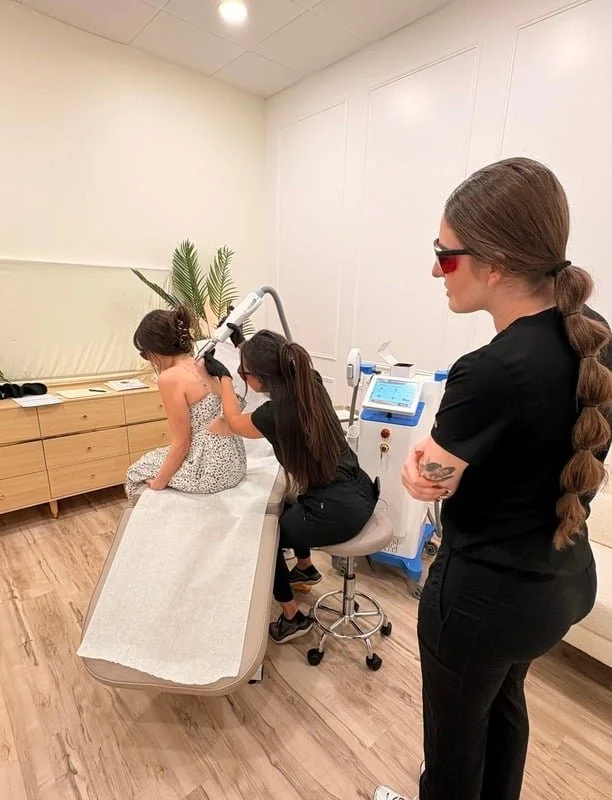 A girl in a polka dot dress is sitting on an examination chair during a laser skincare treatment. A technician is adjusting a laser device near her face, while another woman with long braided brown hair, wearing sunglasses and black scrubs, watches nearby in a clean, modern clinic room.