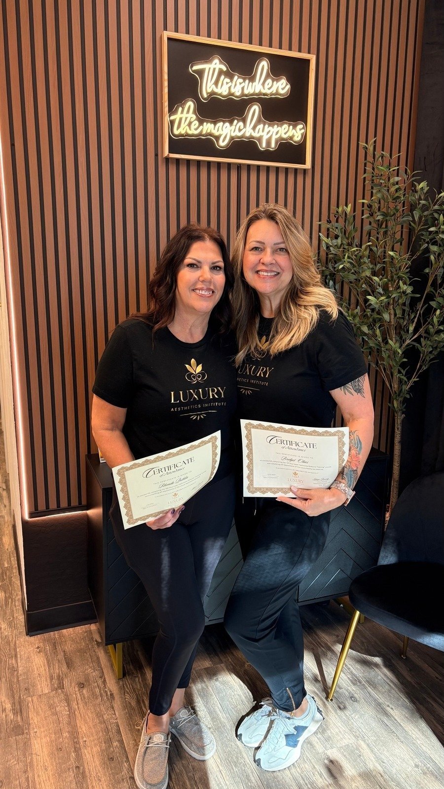Two women holding certificates standing in front of a wooden wall with a neon sign that reads 'This is where the magic happens'. They are smiling and wearing black T-shirts with 'Luxury Aesthetics Institute' logo.