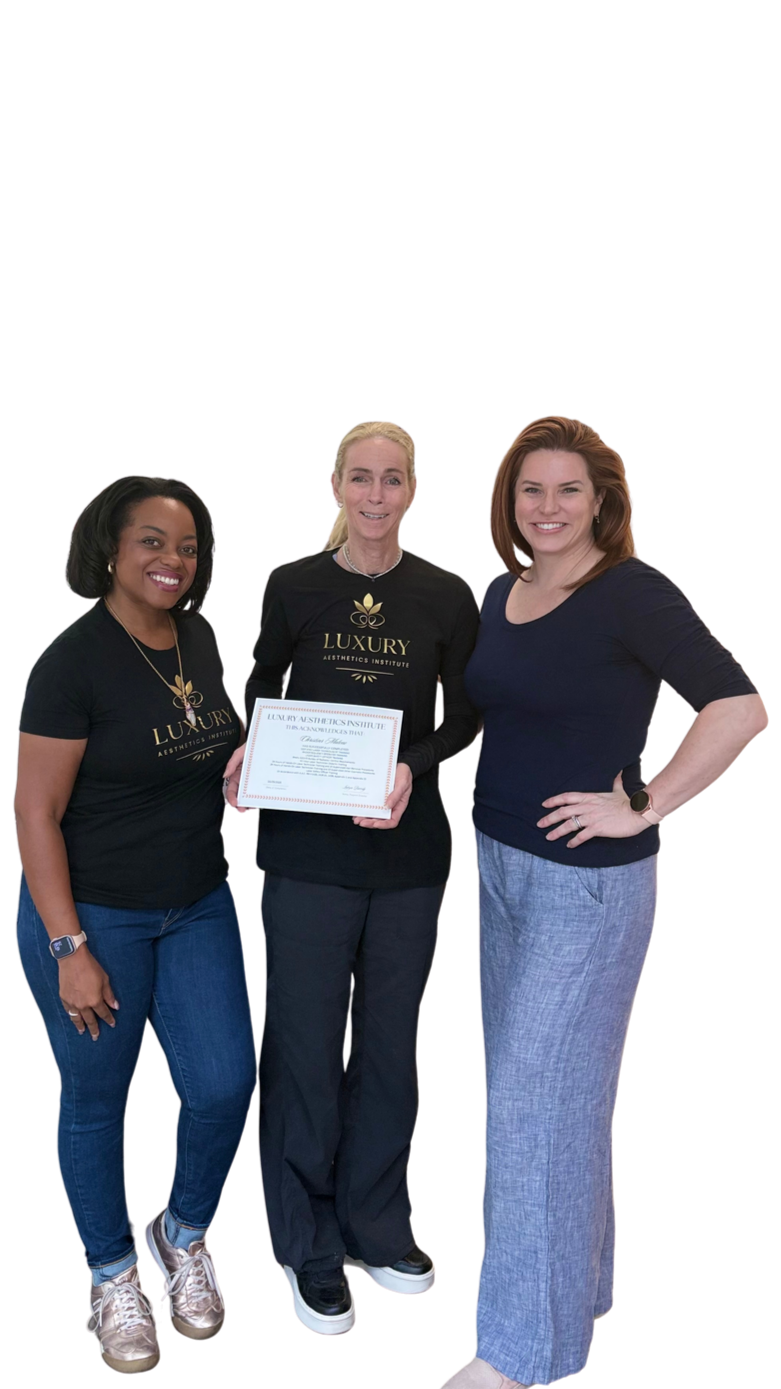Three women standing together, two wearing black T-shirts with a gold logo reading "LUXURY AESTHETICS INSTITUTE," one holding a certificate, smiling at the camera.