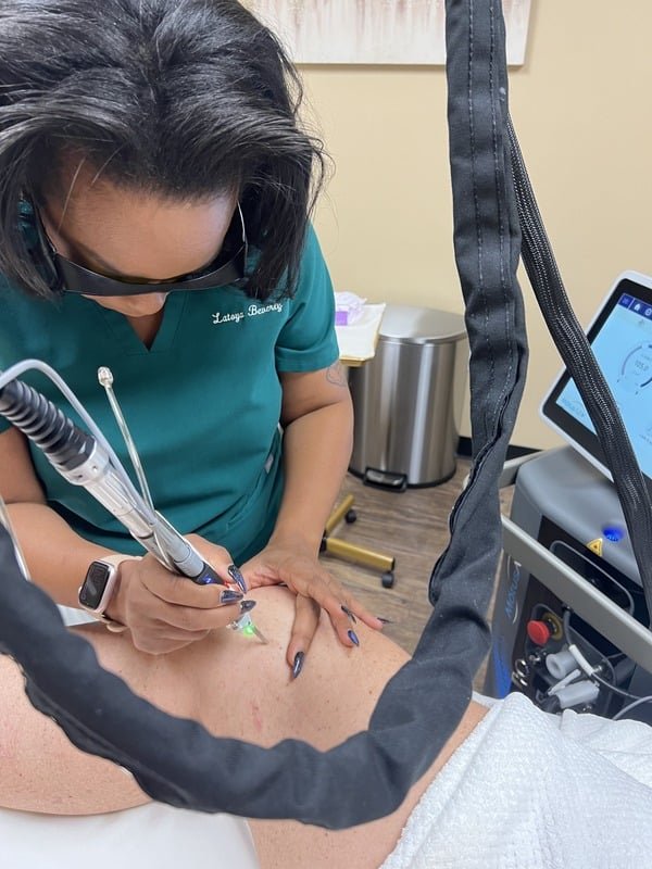A woman in teal scrubs performing a medical procedure on a patient's abdomen in a clinical setting.