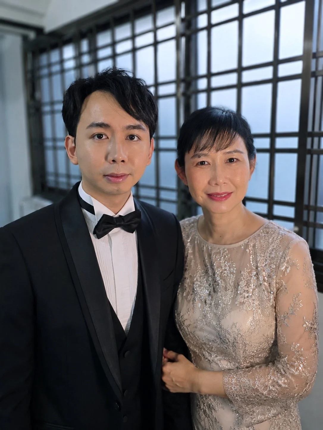A young man in a tuxedo standing next to an older woman in a lace dress, both smiling, with a cityscape background seen through large windows.