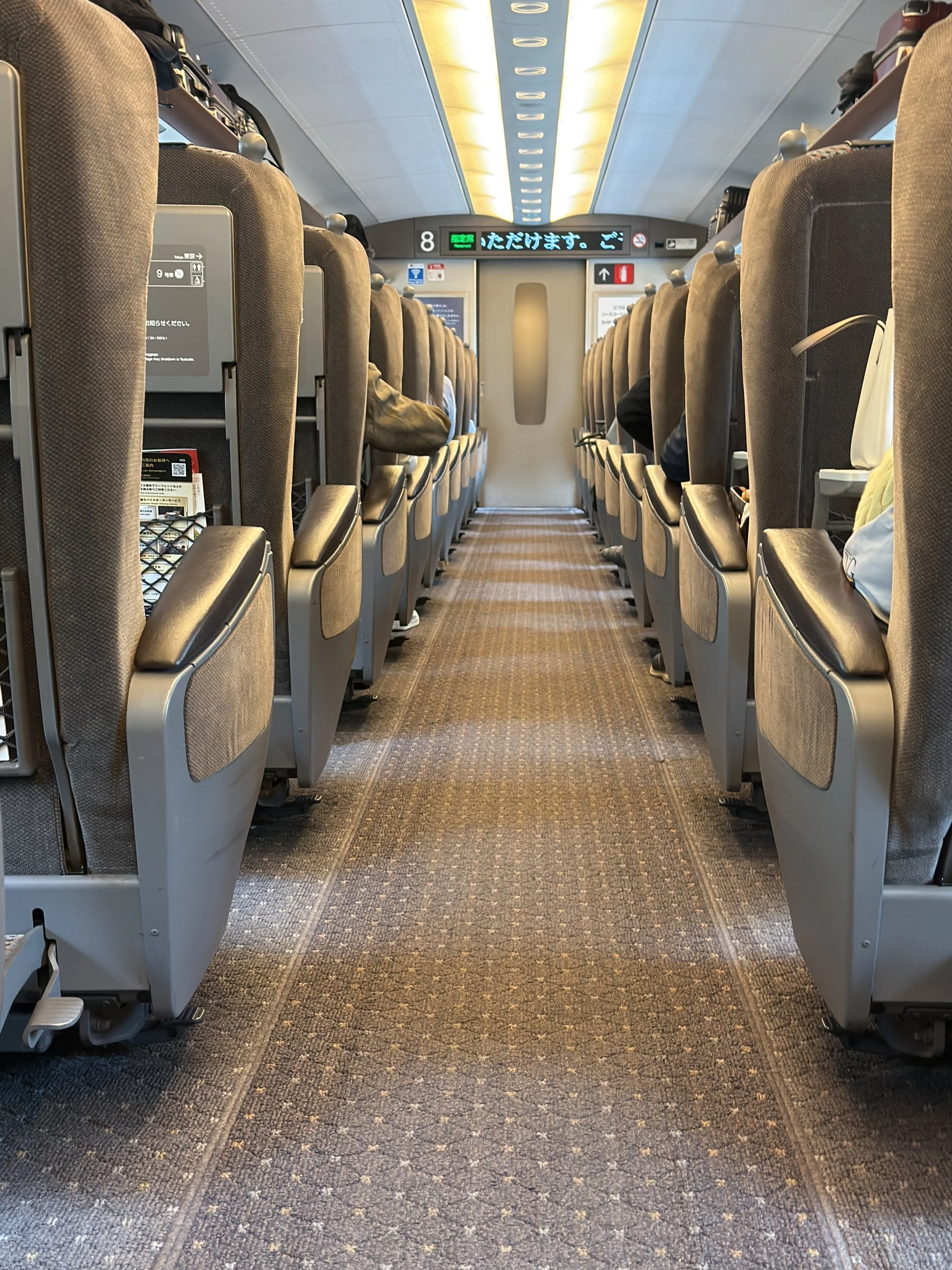 Interior view of an airplane aisle with seats on both sides, some passengers seated, digital signs above the aisle, and a closed door at the end.