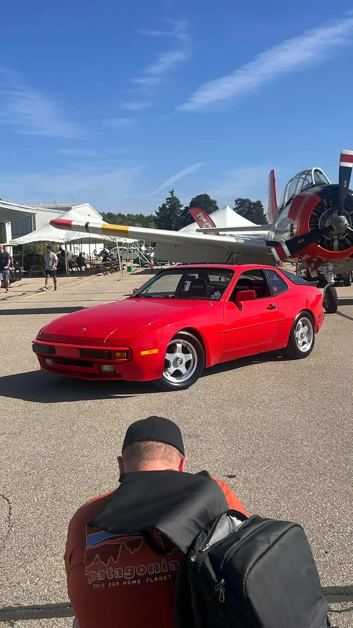 A red sports car parked on a tarmac with a vintage propeller airplane behind it. People are walking around in the background on a clear day.
