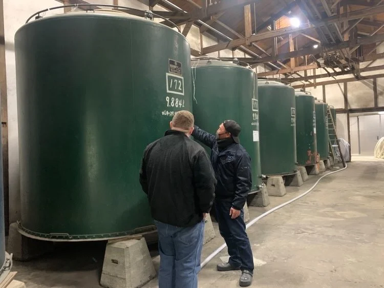 Two men inspecting large green industrial tanks inside a warehouse or factory.