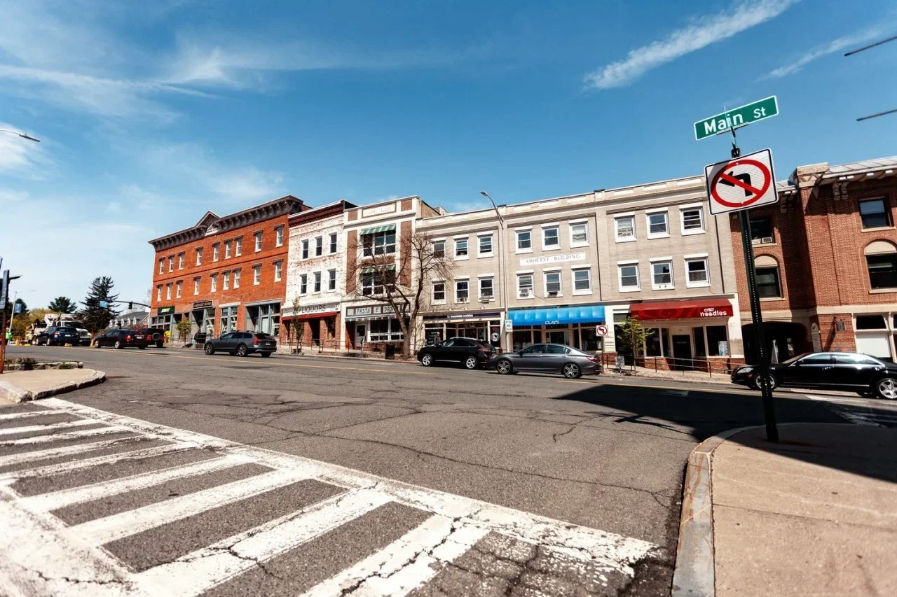 City street with commercial buildings, parked cars, and a no left turn traffic sign under a clear blue sky.