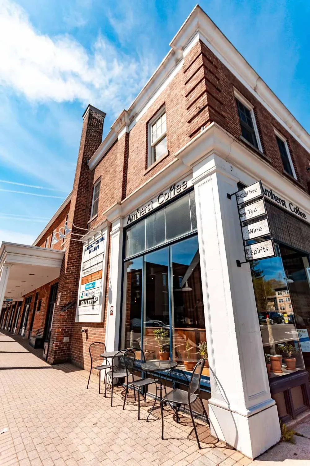 Exterior view of a brick building housing Amherst Coffee with outdoor seating and a sign listing beverages like tea, wine, and spirits on a bright, sunny day.