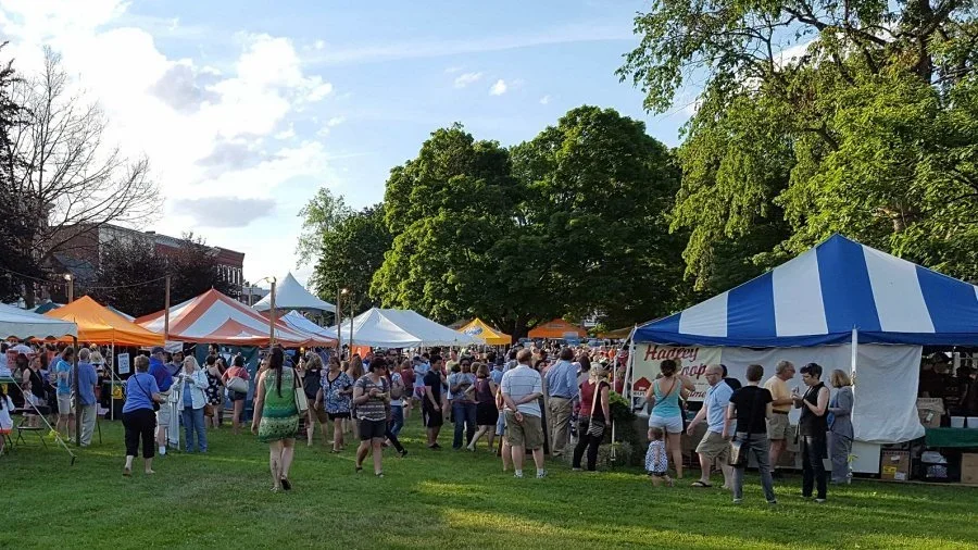 People walking and shopping at an outdoor fair with multiple tents and food stalls, greenery, and trees in the background, under a partly cloudy sky.