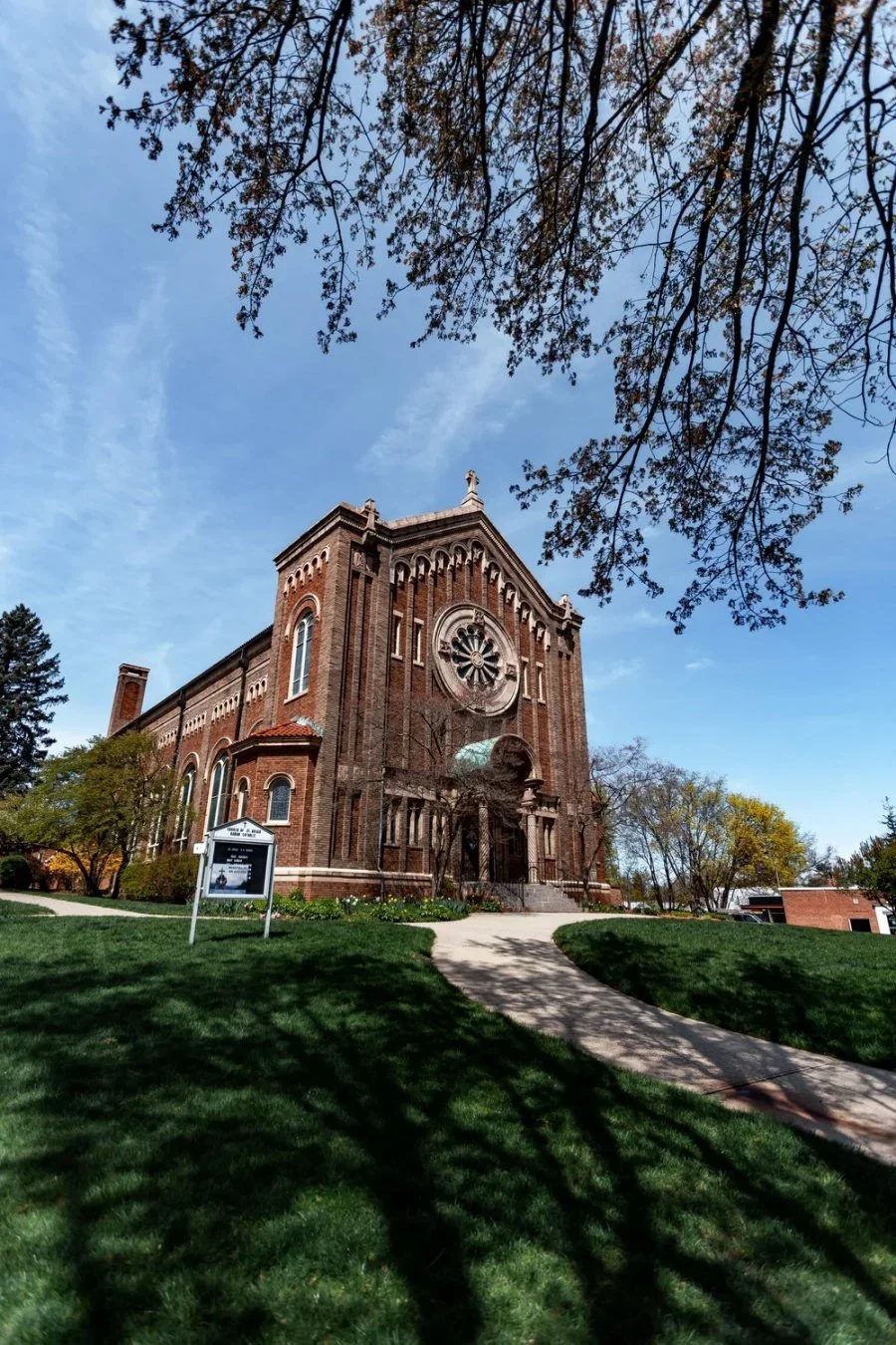 A tall brick church with a large circular stained glass window in the front, surrounded by a well-maintained green lawn with a sidewalk leading to the entrance, and trees with some leaves in fall colors under a blue sky.