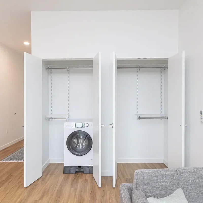 An empty white closet with a washing machine inside on the left side, and space for clothes on either side. The closet has open doors, and the floor is wooden.