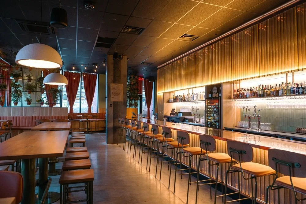 Interior of a modern bar with a long counter, various bottles of alcohol, bar stools, hanging pendant lights, and windows with red curtains.