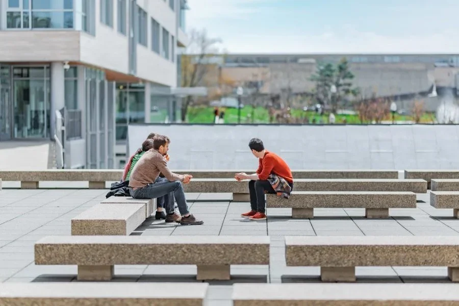 Three people sitting on concrete benches outside, engaged in conversation. They are in an urban setting with modern buildings and a green park in the background.