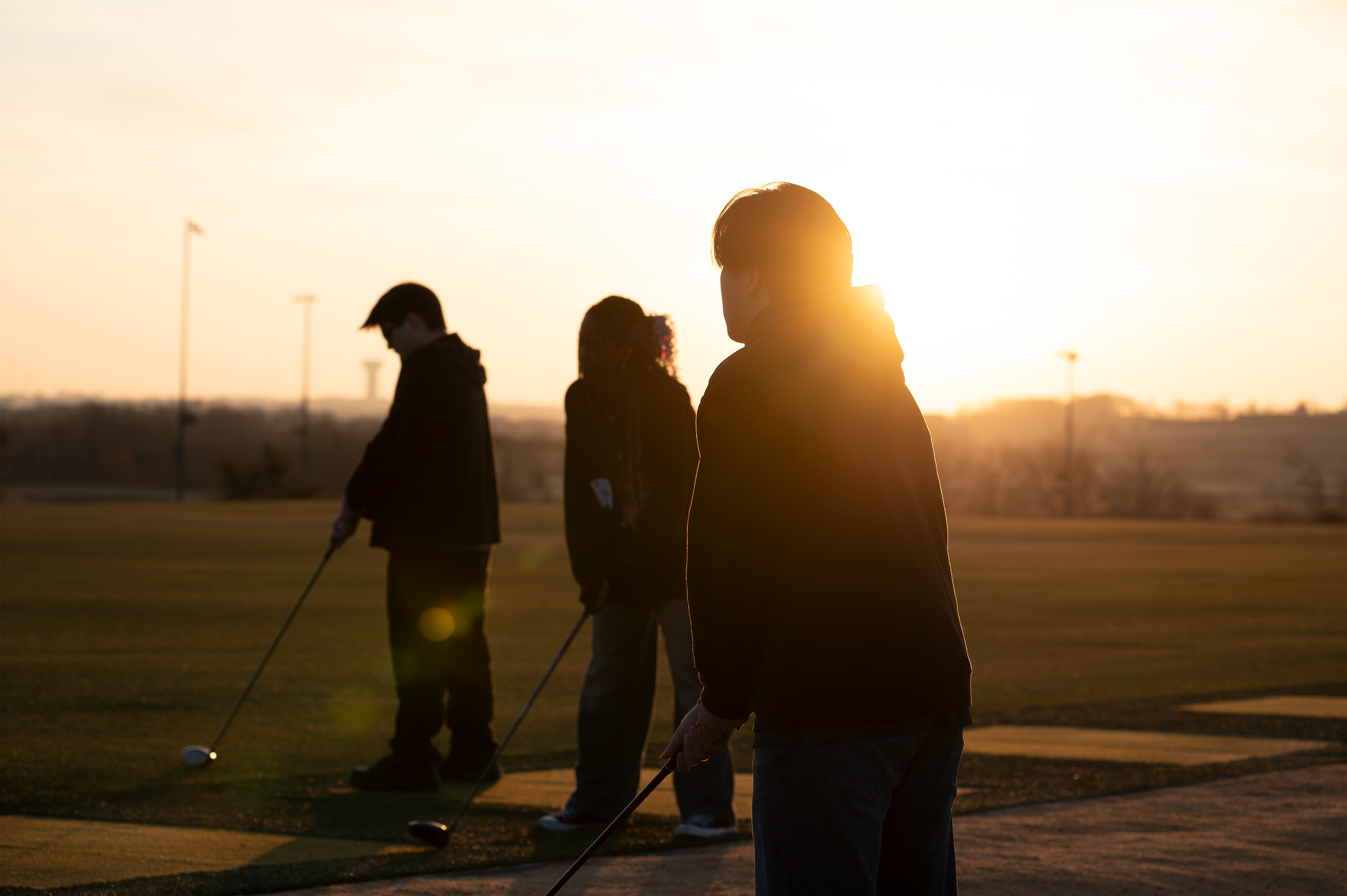 Silhouettes of three people playing golf on a course during sunset.