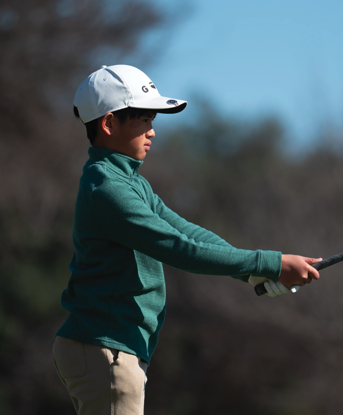 Young boy wearing a white cap and teal jacket, holding a golf club on a golf course with a blurred natural background.