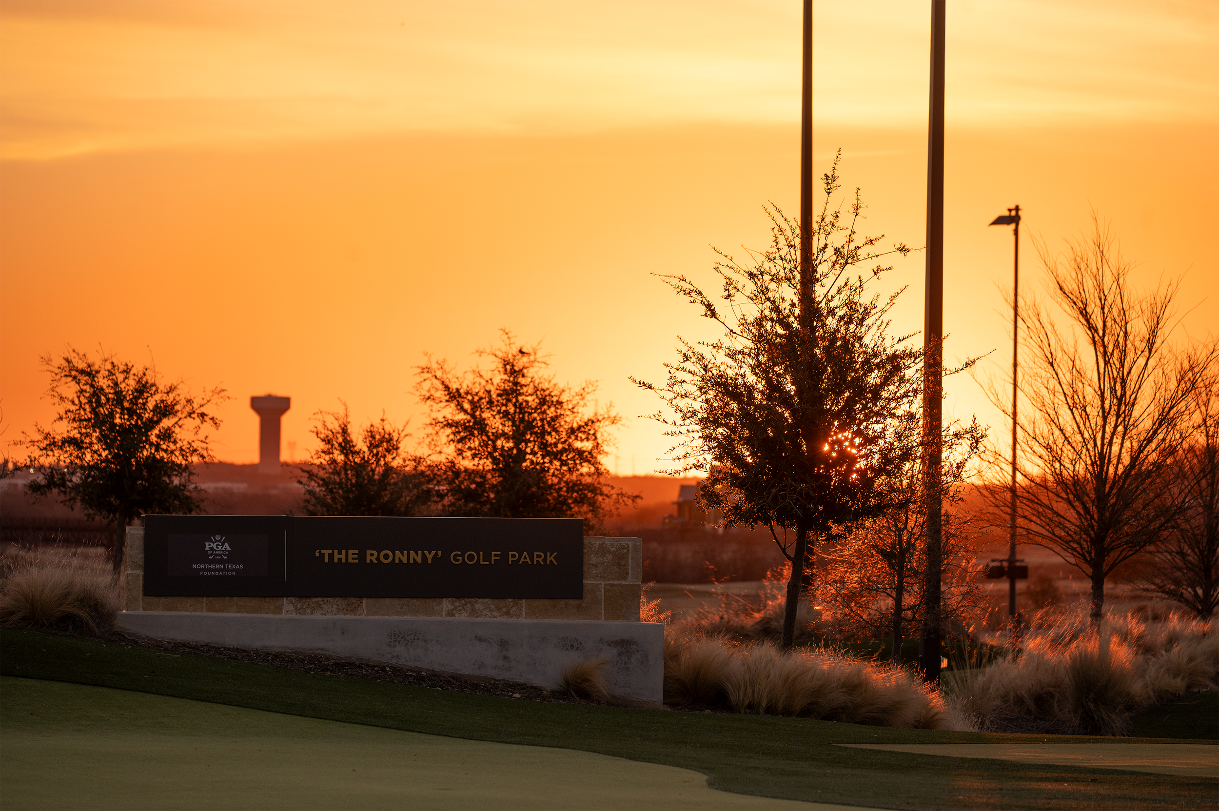 Sunset over The Ronny Golf Park with a sign, trees, and light poles silhouetted against an orange sky.
