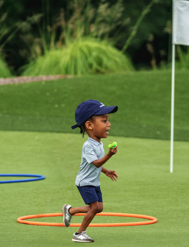 Young boy running on a green field with hula hoops, holding a small green ball.