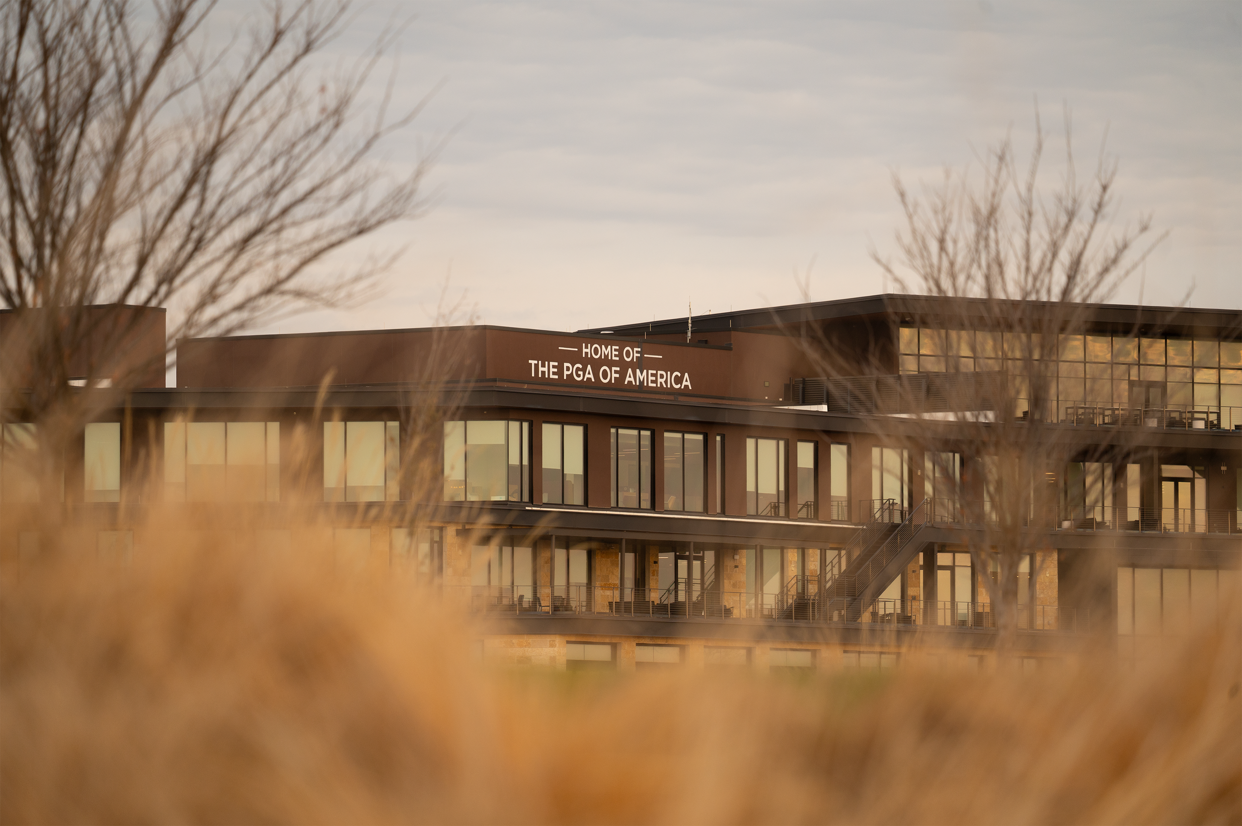 A modern building with large glass windows, a sign that reads "Home of the PGA of America," and a staircase on the exterior, seen through some blurred grasses and trees in the foreground.