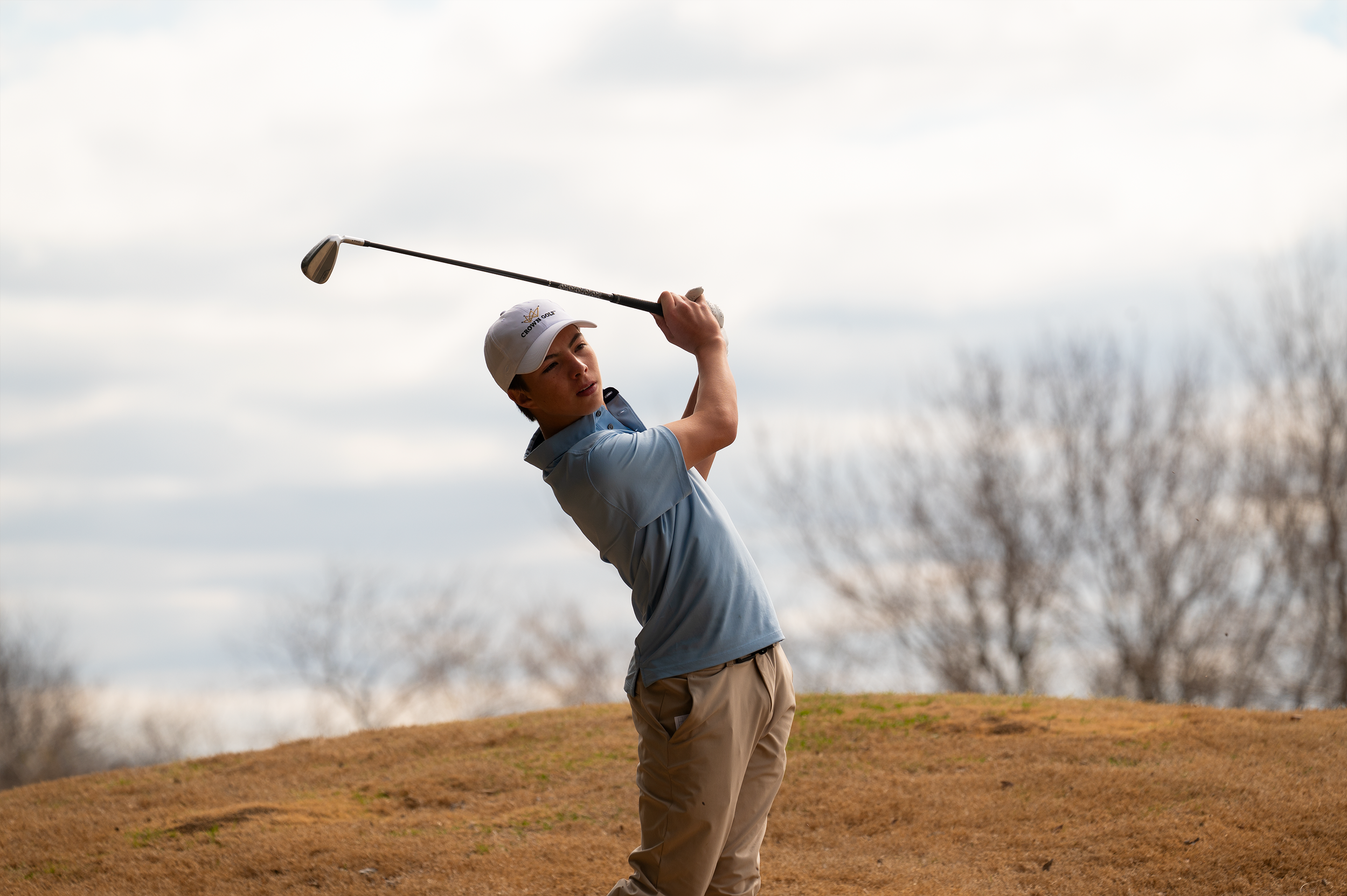 Young man in a blue polo and tan pants taking a golf swing on a golf course with trees and cloudy sky in background.