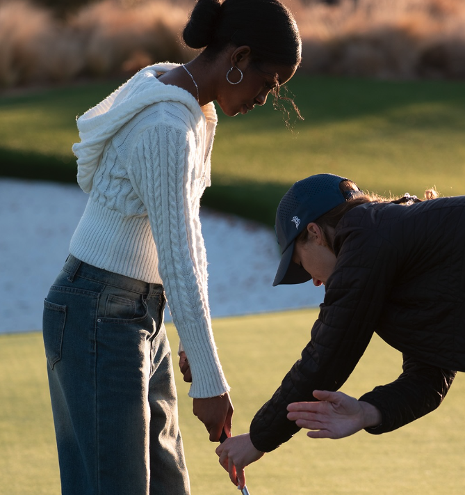 Two women preparing to putt on a golf course, one helping the other.