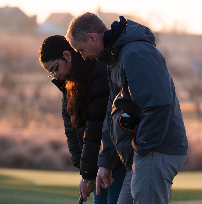 A man and woman standing closely on a golf course at sunset, both dressed in jackets, with their heads bowed and eyes closed.