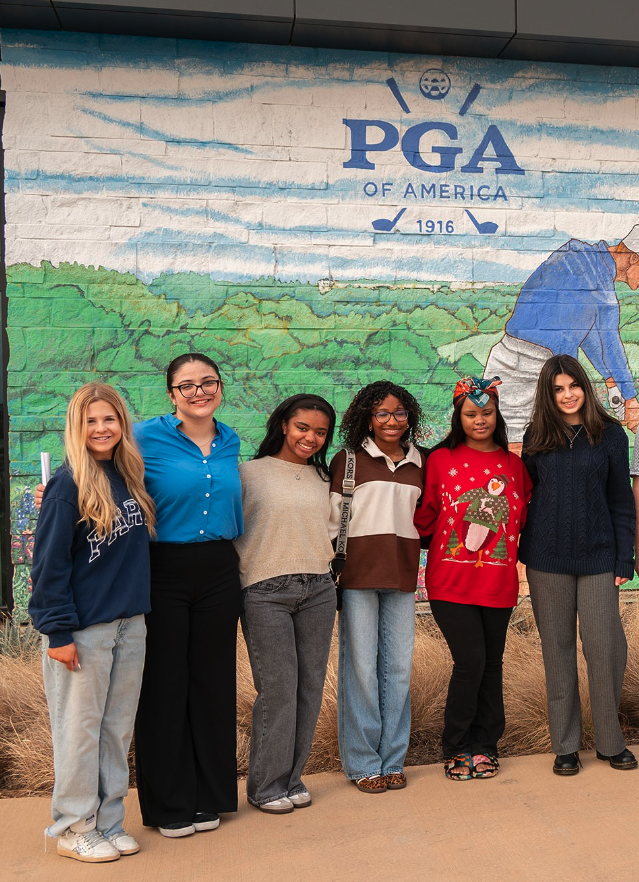 Group of six young women standing in front of a mural with the PGA of America logo, trees, and grass, smiling at the camera.