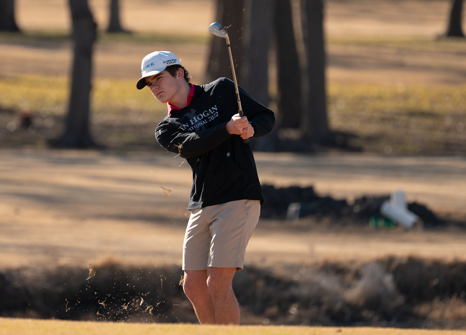 Young male golfer taking a shot from a sand bunker on a golf course with trees in the background.