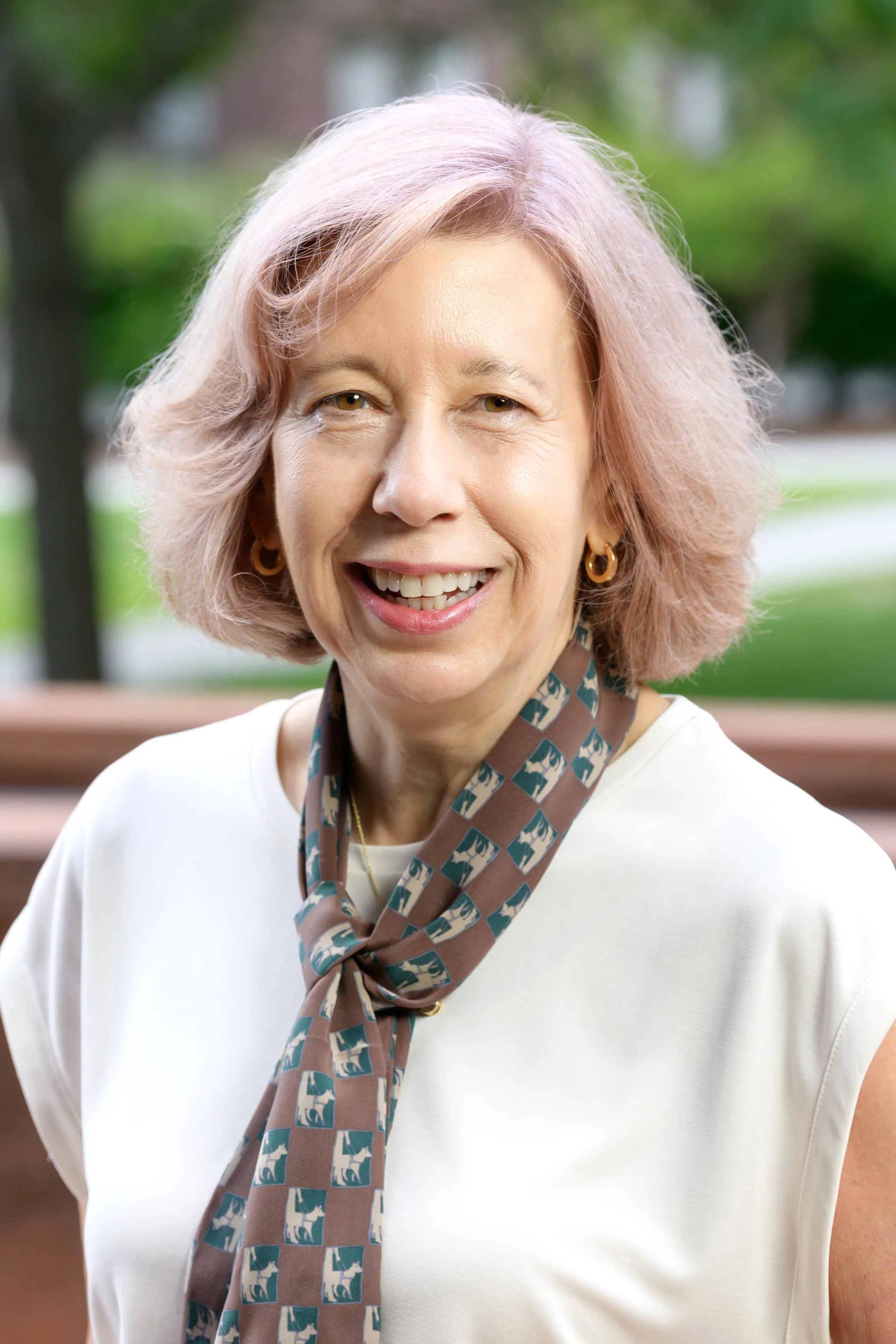 A smiling middle-aged woman with light pink hair outside, wearing a white top and a patterned scarf, blurred green background.