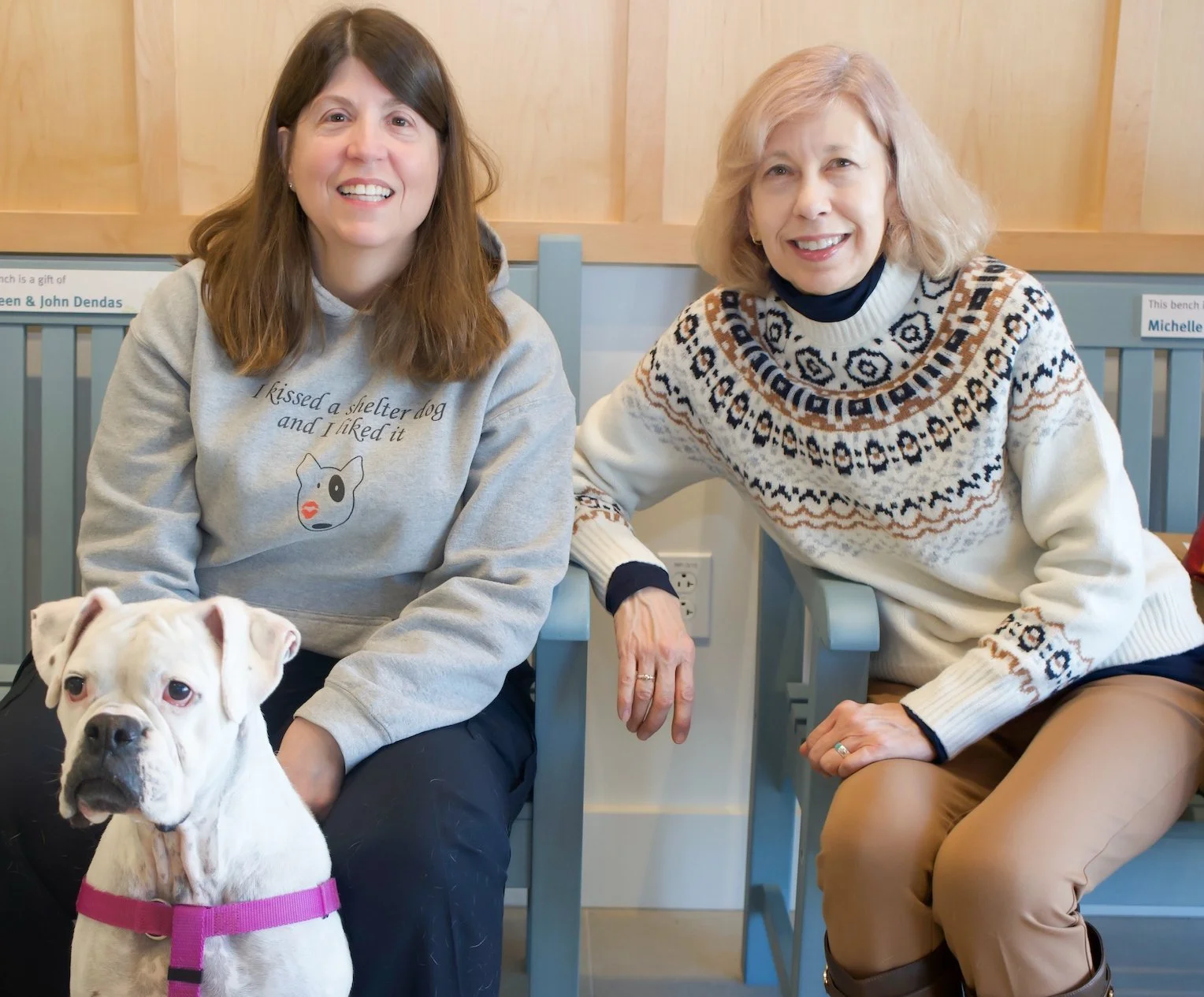 Two women sitting on chairs with a white dog in front of them. The woman on the left has long brown hair, is smiling, and is wearing a gray sweatshirt with a dog illustration and text. The woman on the right has blond hair, is smiling, and is wearing a cream-colored sweater with a patterned design. The dog is wearing a pink collar.