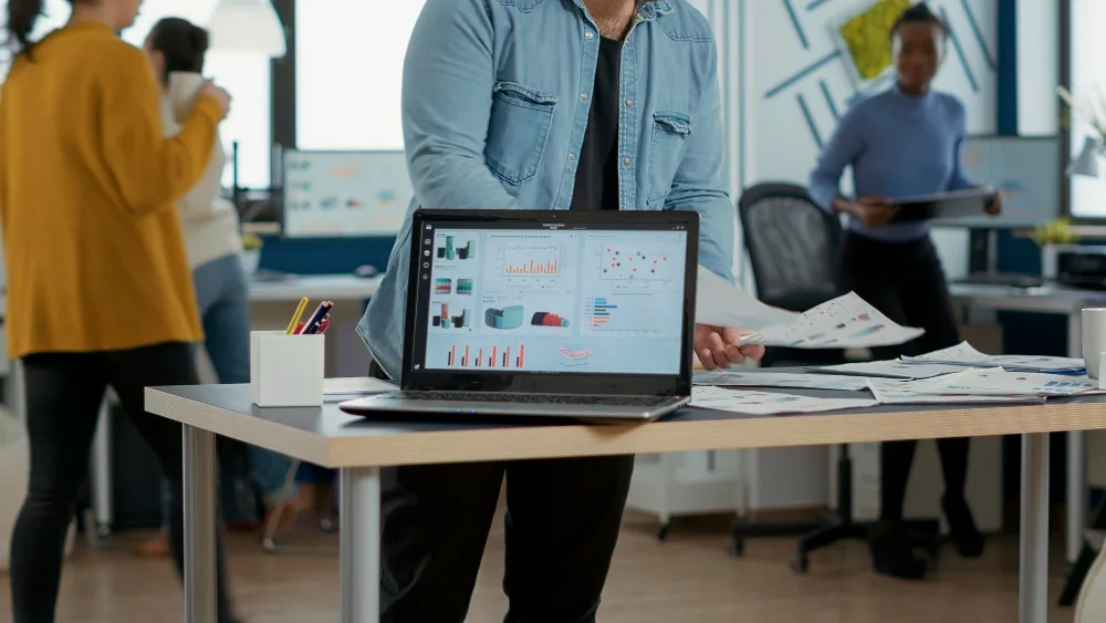 A person standing at a desk in a modern office, holding a sheet of paper, with a laptop displaying graphs and charts. Other colleagues are working in the background.