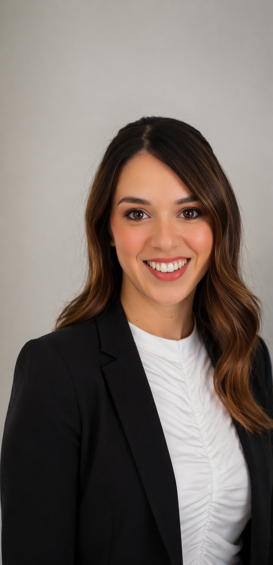 A professional woman with long, wavy brown hair, smiling, wearing a black blazer and a white blouse, standing against a plain light gray background.