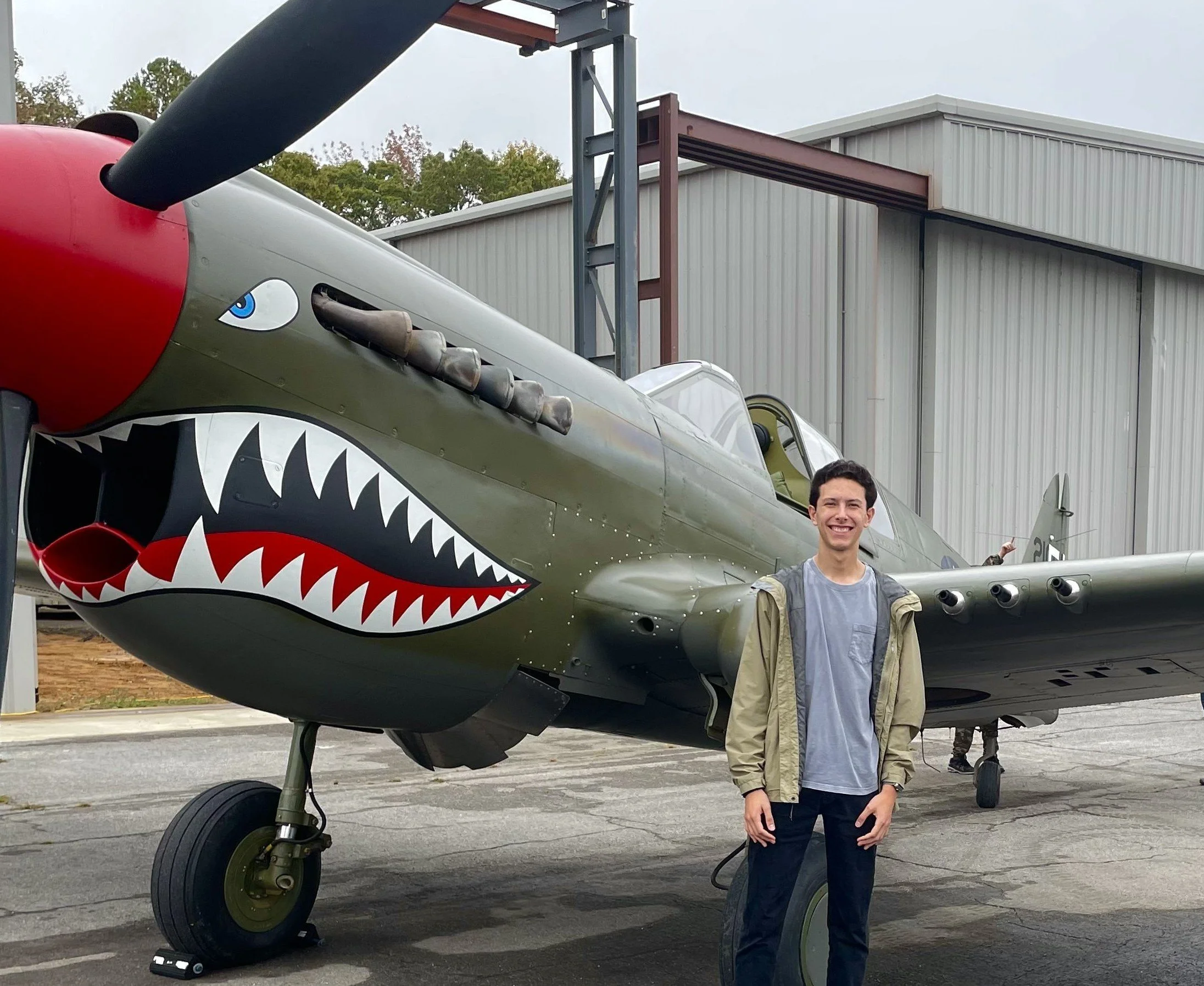 A young man standing in front of a vintage military aircraft with a shark mouth painted on the nose, outside a hangar on an overcast day.