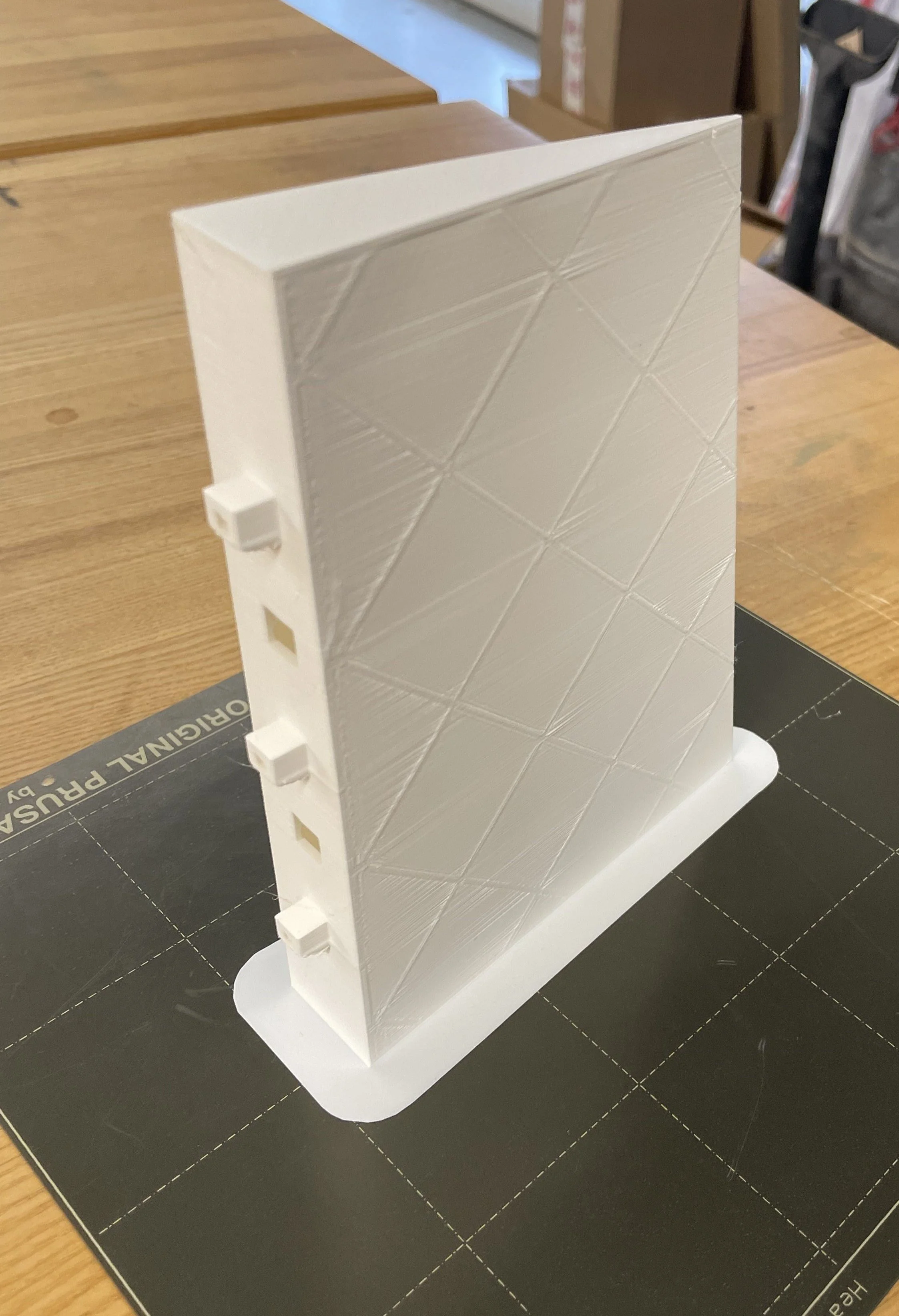 3D-printed white model of a building with a textured diamond pattern on the facade, placed on a black cutting mat on a wooden table.