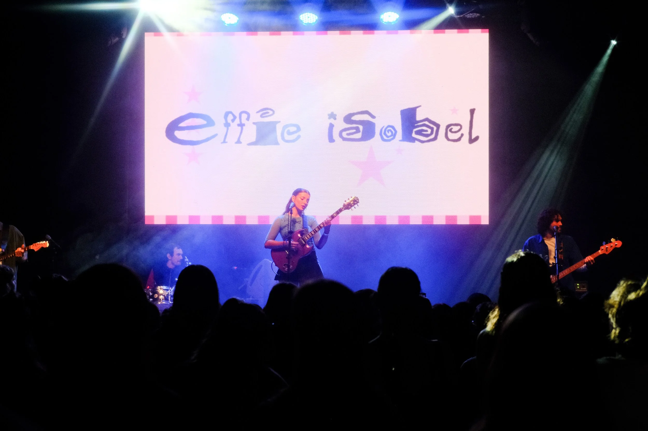 A band performs on stage at a concert with a woman playing an acoustic guitar at center, a drummer behind her, and two other band members playing guitars. A large screen behind displays the text 'Effie Isabel' with pink stars around it, amid purple and blue stage lighting.