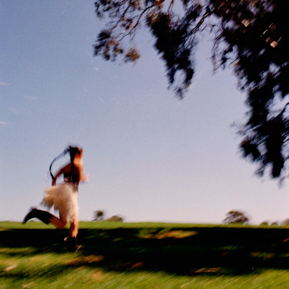 A person in a white dress running on a grassy area, with trees and a blue sky in the background, in motion blur.