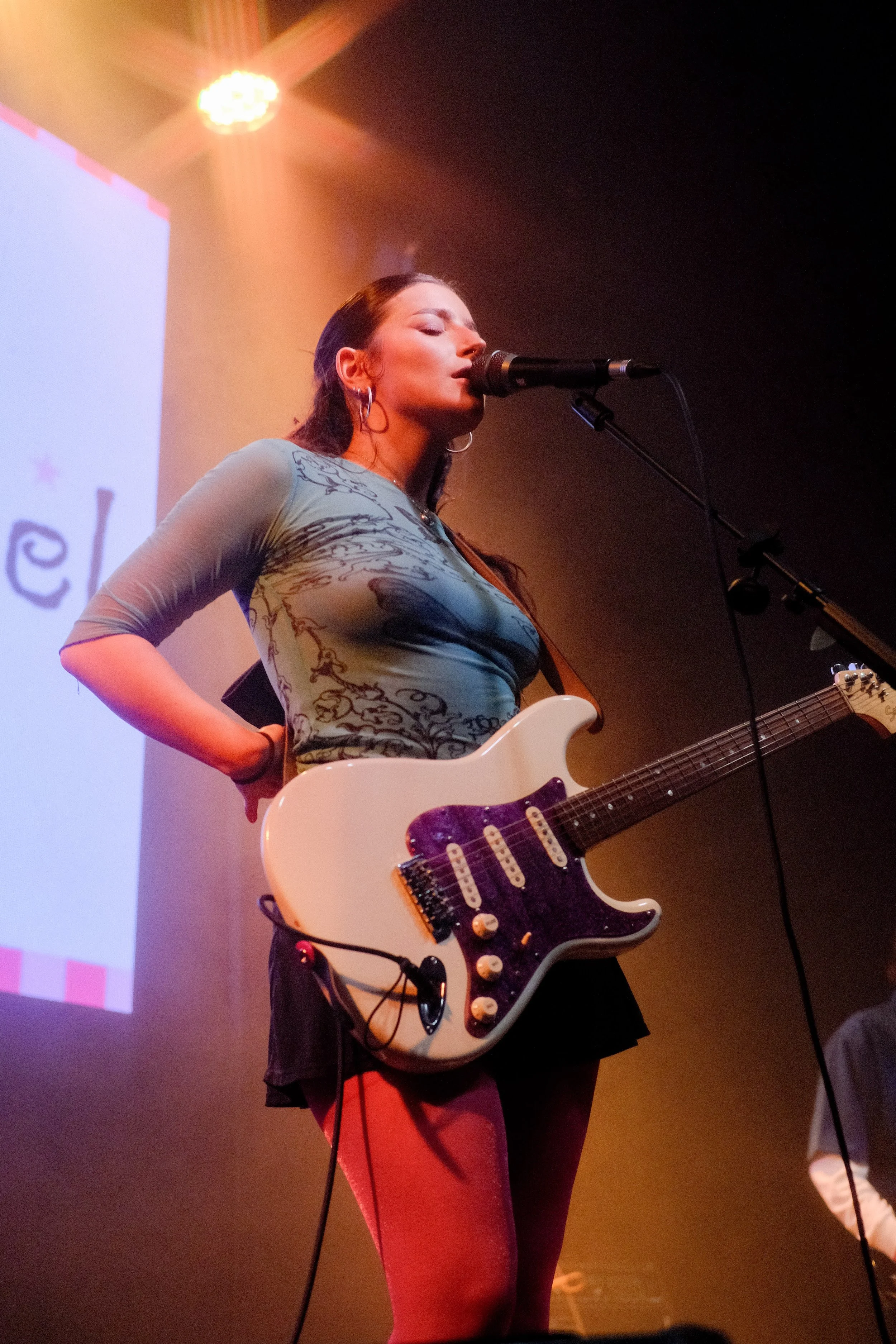 A woman with long dark hair singing into a microphone while holding an electric guitar on stage, with stage lighting and a screen in the background.