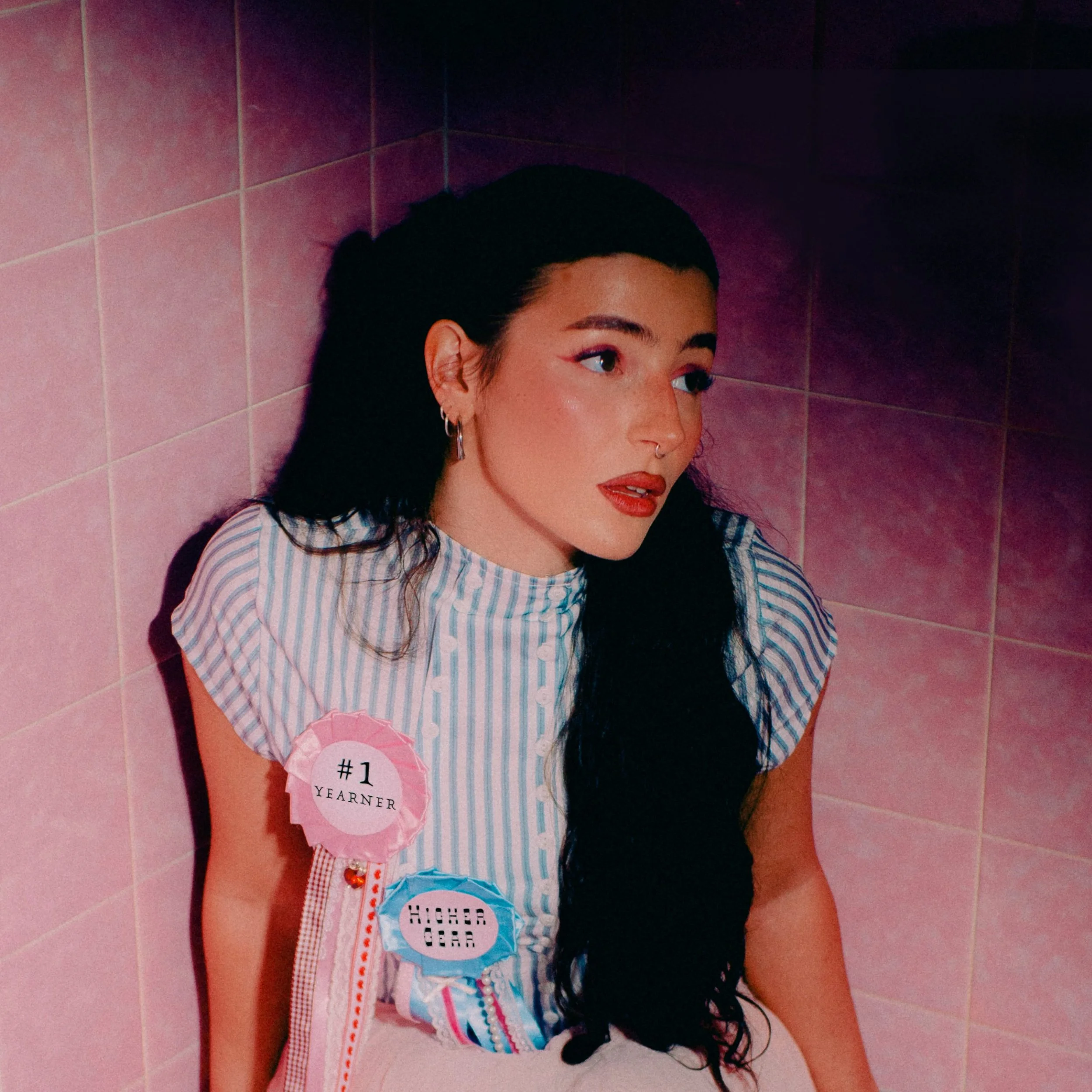 A young woman with long black hair, wearing a striped blue and white top, is leaning against pink tiled wall. She has earrings, a septum piercing, and makeup with bold red lipstick. She has two badges pinned to her top: one saying '#1 Yearner' and the other 'Higher Gears'.