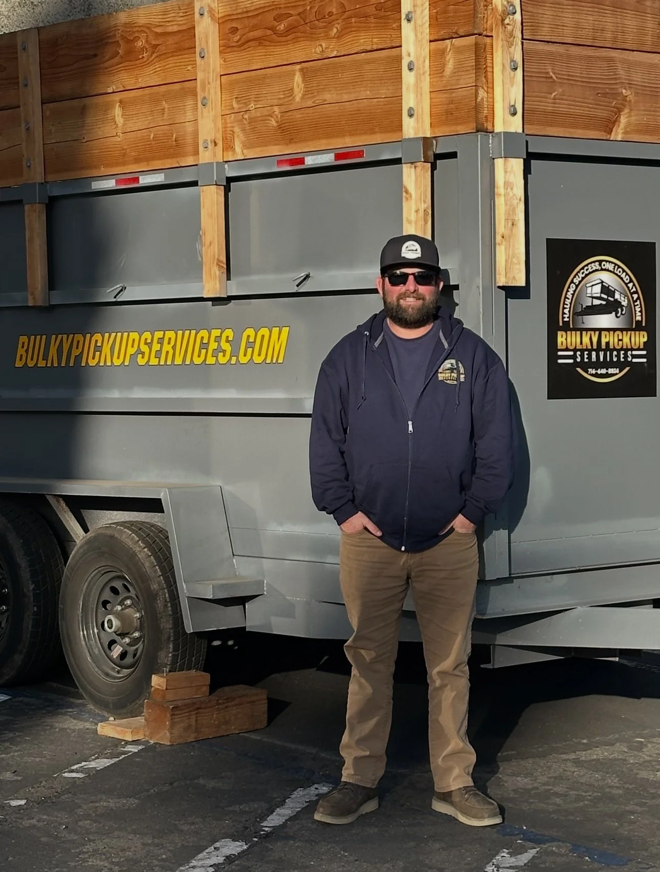 A man standing in front of a company vehicle with a logo that reads 'Bulky Pickup Services,' wearing sunglasses, a hoodie, and beige pants, with wood being transported in the truck bed.