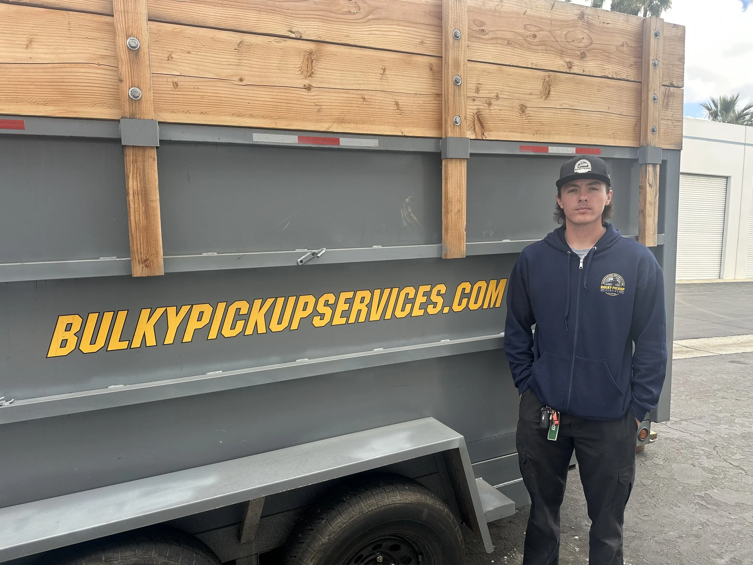 Young man standing outside next to a large truck with wooden panels on top and the website 'BULKY PICKUP SERVICES.COM' written on the side.