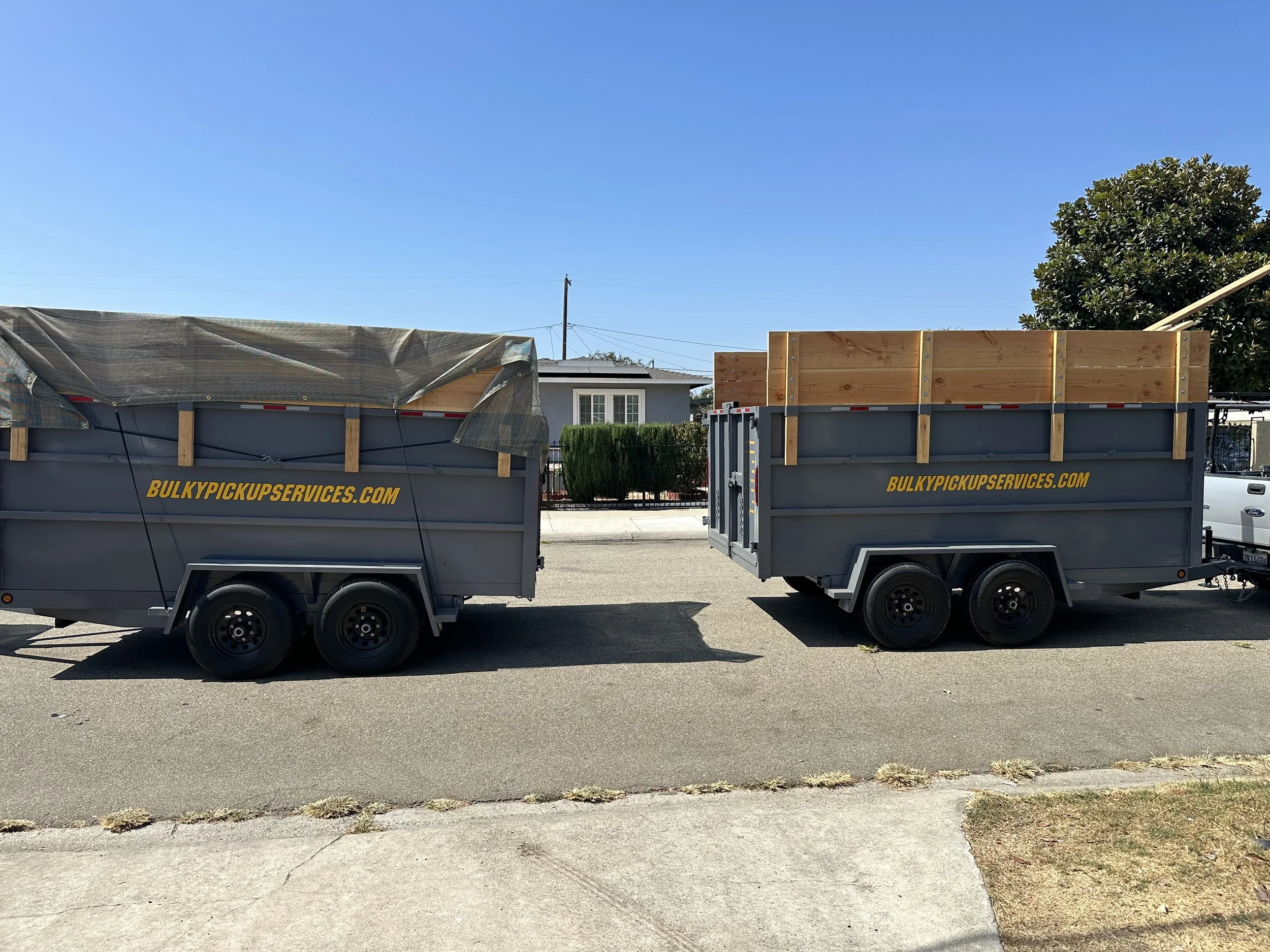 Two grey utility trailers parked side by side on a street, with wooden frames in the trailers, and the website 'BULKYPICKUPSERVICES.COM' written on the sides.