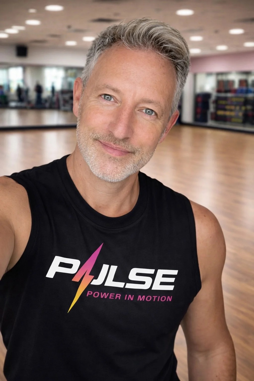 A smiling, middle-aged man with gray hair and a beard taking a selfie in a gym, wearing a black sleeveless shirt with the logo 'PULSE Power in Motion' on it.