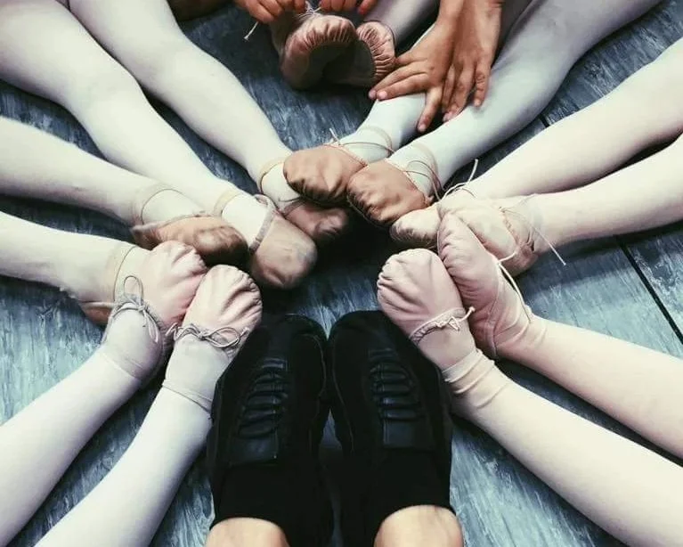 Ballet dancers in ballet slippers forming a circle with their feet, with an instructor's hand in the middle, on a wooden floor.