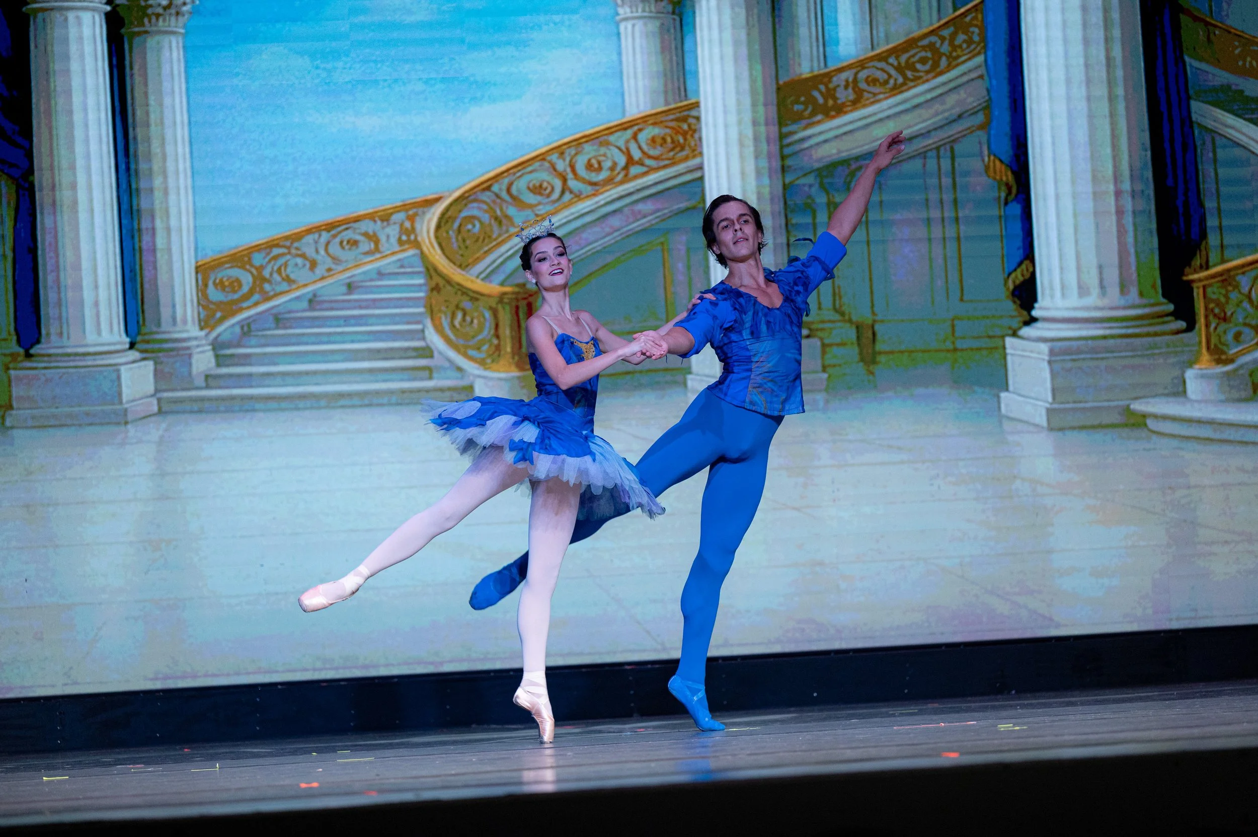 Two ballet dancers performing on stage with a backdrop of a painted staircase and ornate columns, dressed in blue costumes.