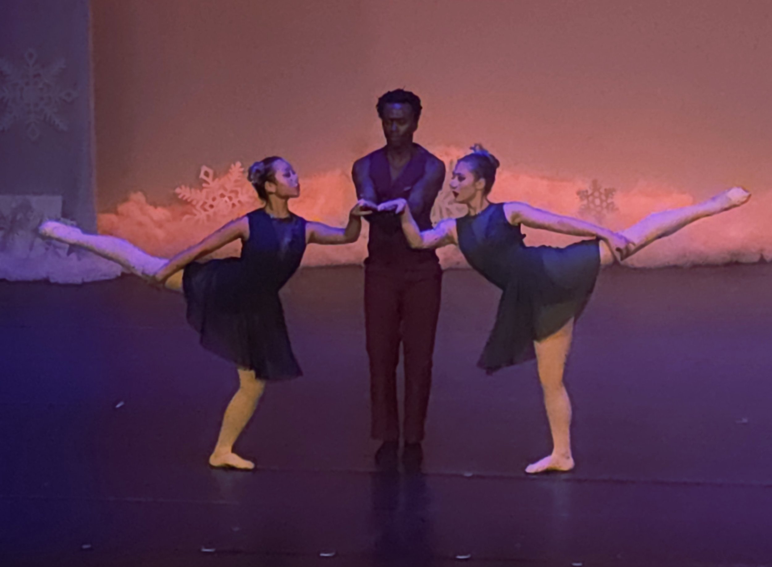 Two ballet dancers in black leotards and yellow tights performing with a male dancer in a black shirt and pants on stage with pink and purple lighting and snowflake decorations in the background.