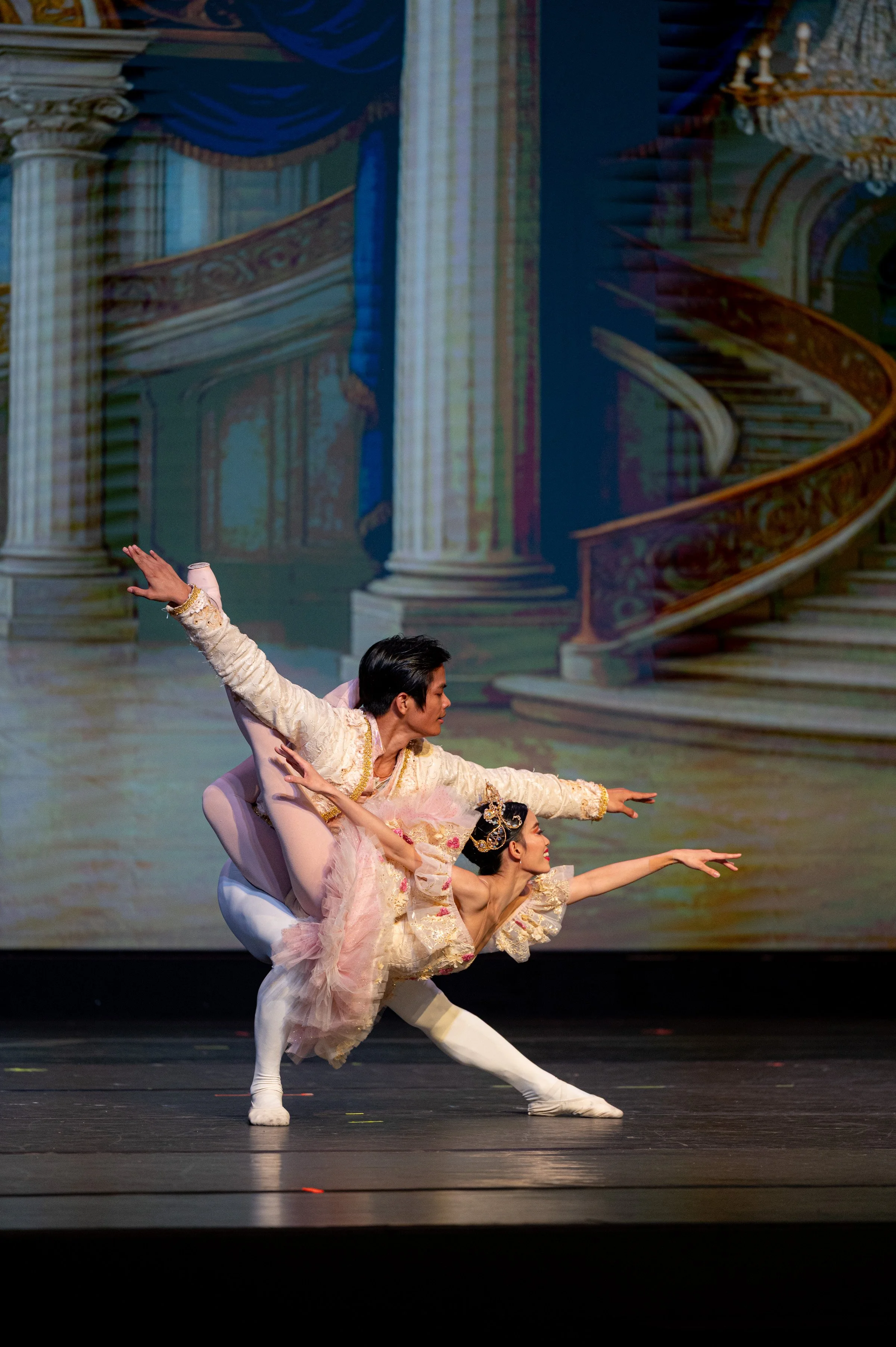 Ballet dancers performing on stage with a backdrop of a grand staircase and columns from a palace or theater, dressed in elaborate costumes.