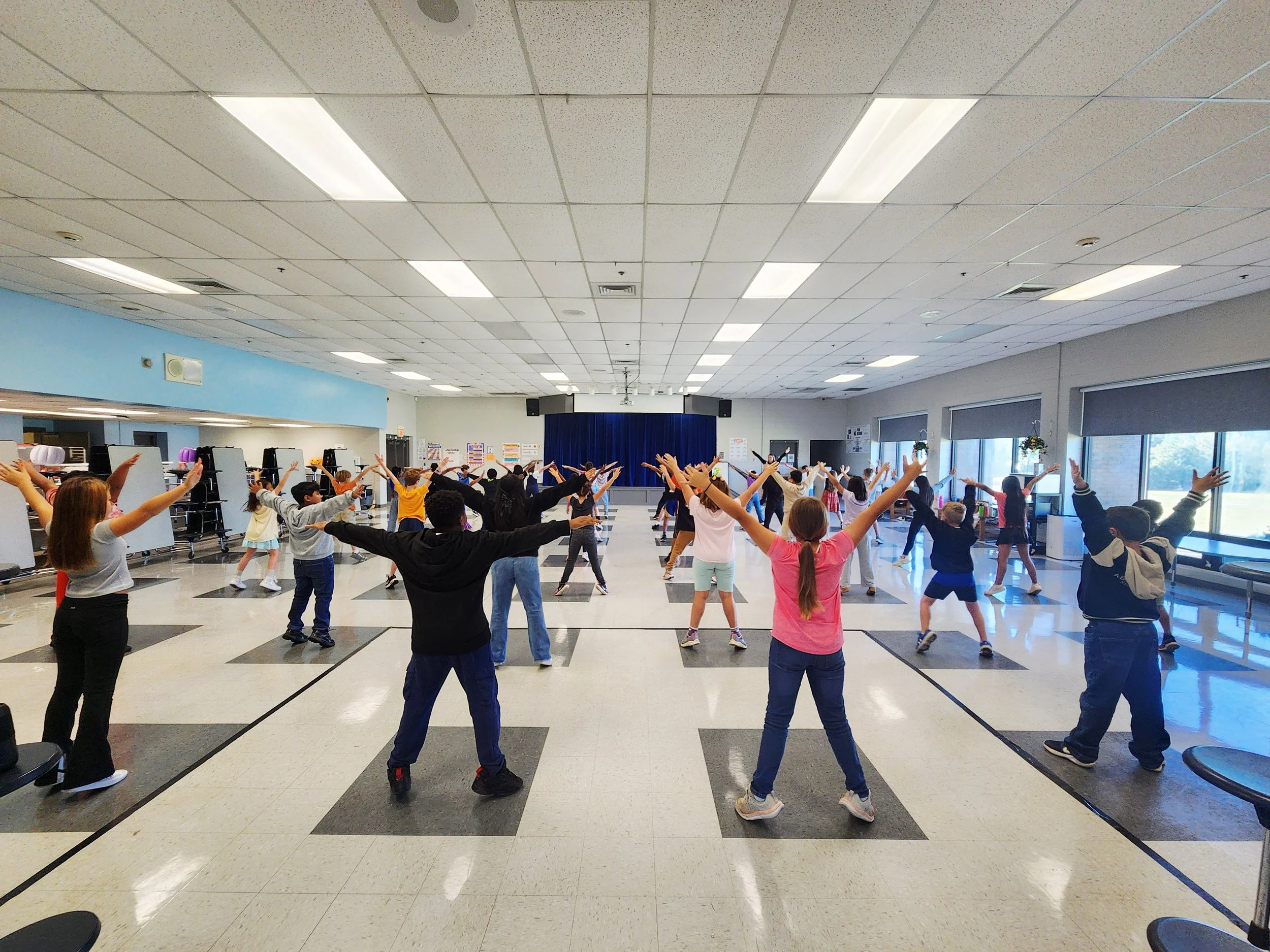 A large group of children participating in a dance or exercise class in a spacious room with a high ceiling, fluorescent lights, and large windows on the right side. The children are spread out evenly and are following a leader or instructor.