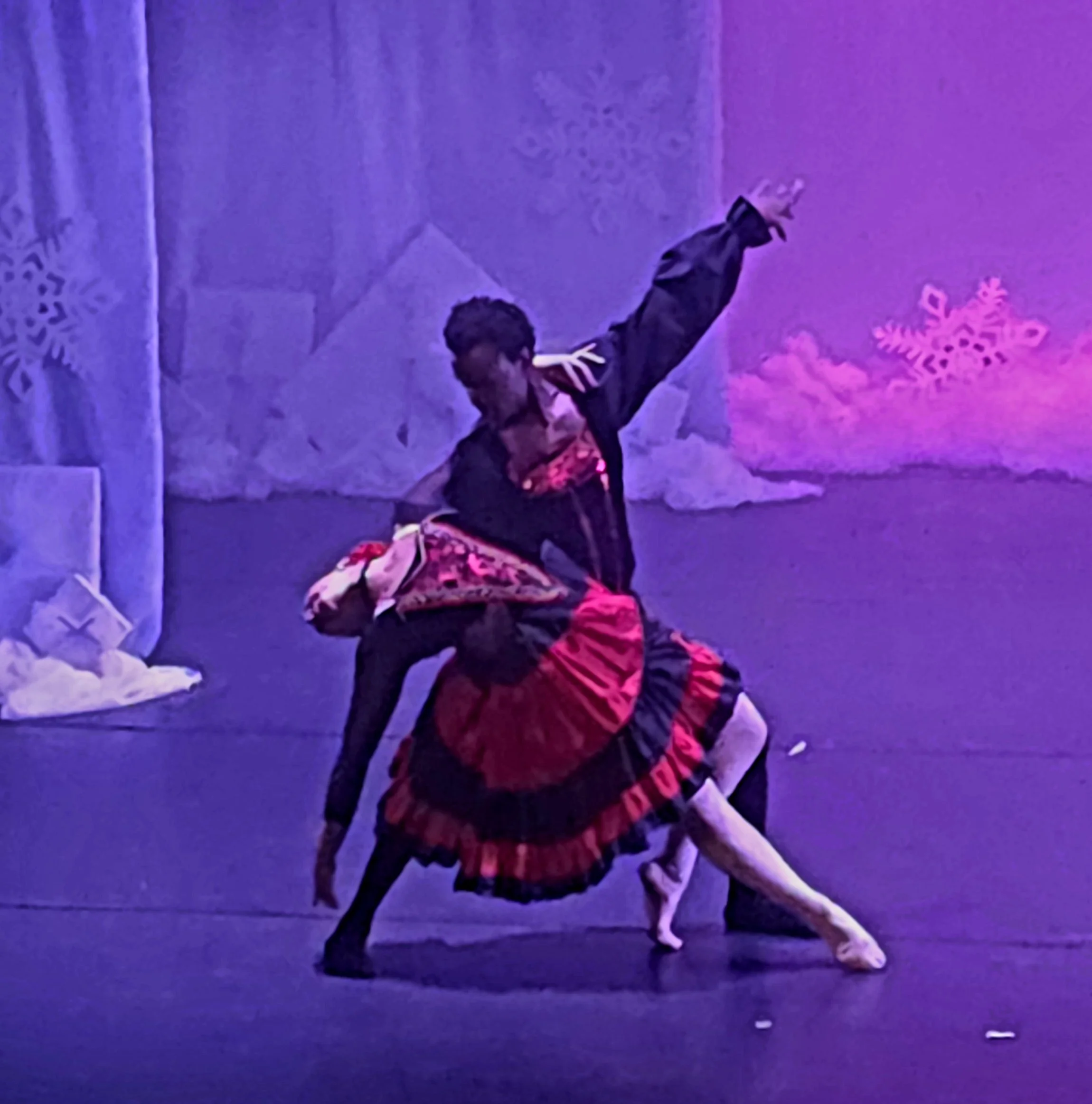 Two children perform a ballet dance on a stage decorated with snowflakes and star cutouts, bathed in purple and pink lighting.