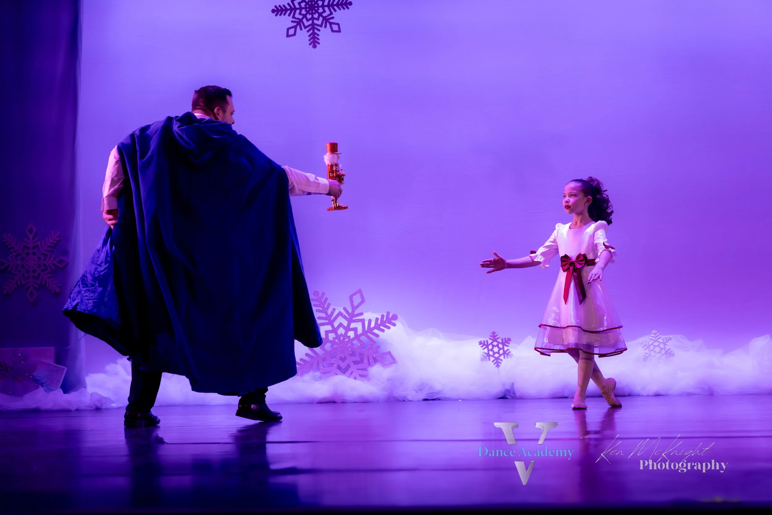 A dance performance on stage featuring a man in a dark cape and a young girl in a white dress with a red bow, with snowflake decorations and purple lighting.